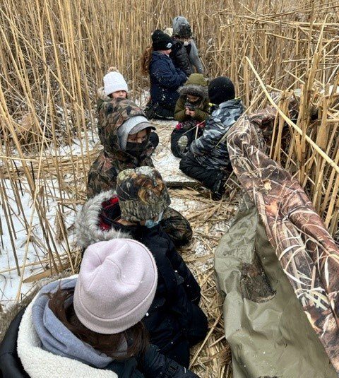 Students participating in a land-based learning activity in winter wetlands, sitting together among tall reeds.